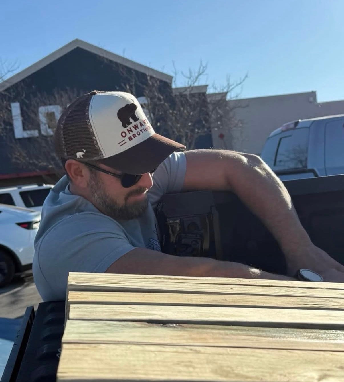 Man in a truck bed with wooden planks, wearing a cap and sunglasses.