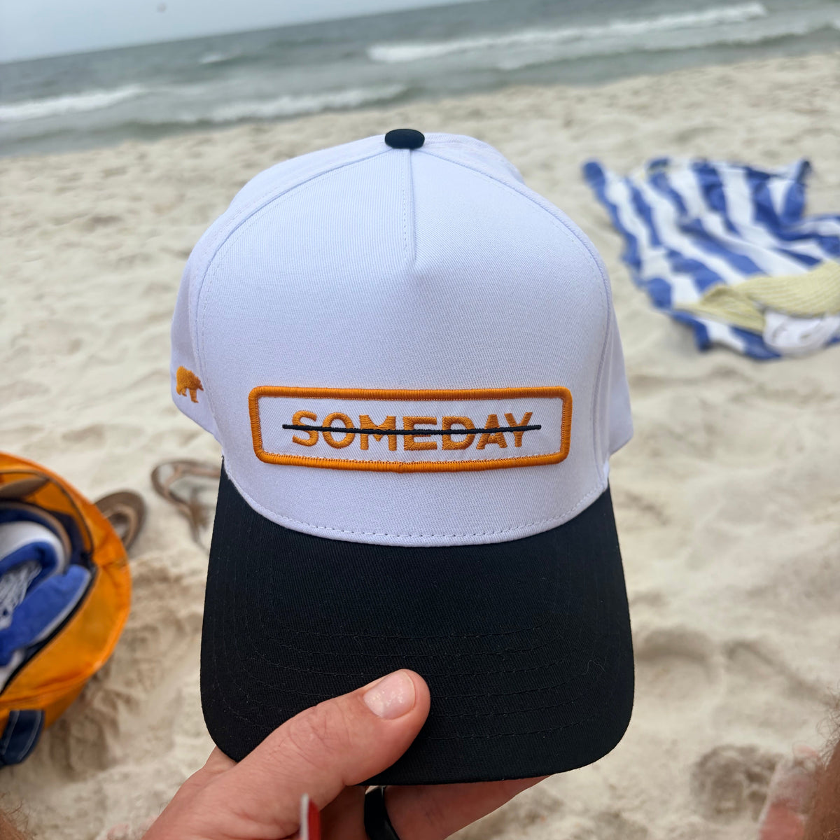 A white and black baseball cap with an orange embroidered 'SOMEDAY' patch on the front, held by a hand on a sandy beach with the ocean in the background.