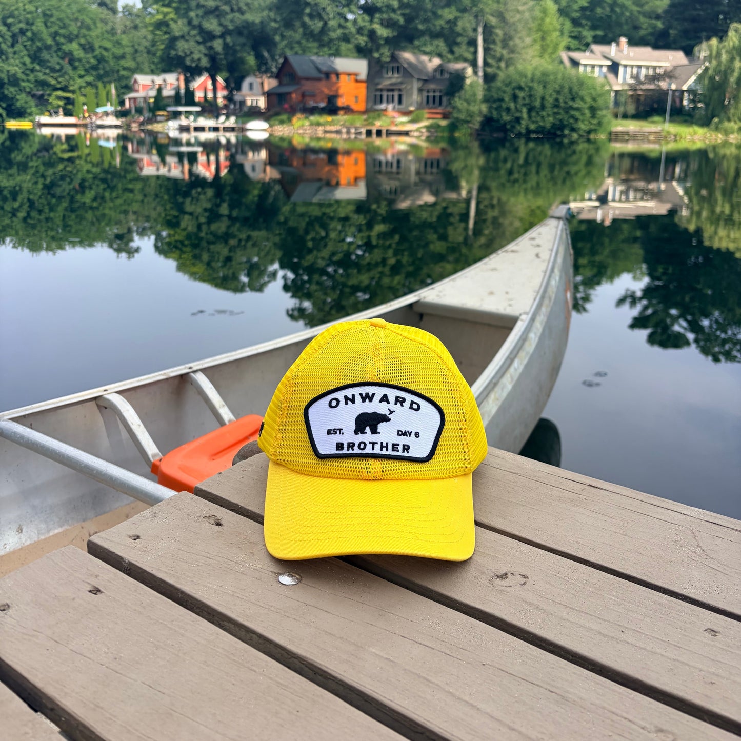 A bright yellow trucker hat with a black and white 'ONWARD EST.  day 6 BROTHER' patch sits on a wooden dock next to a canoe, with a calm lake, trees, and house reflections in the background.