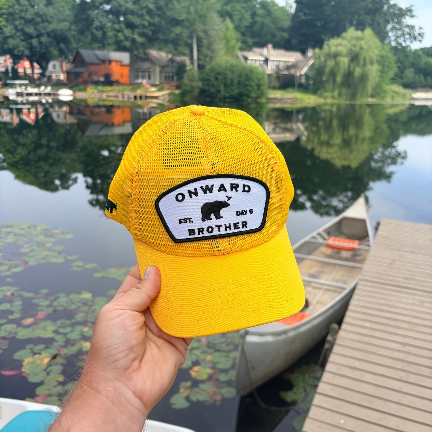 A bright yellow trucker hat held up by a hand, featuring a black and white 'ONWARD EST. day 6 BROTHER' patch with a bear logo.