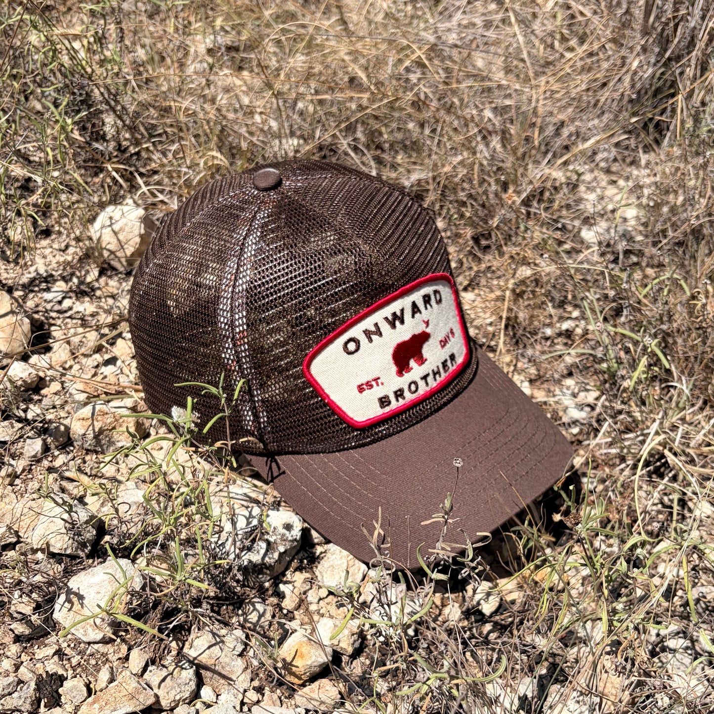 A brown trucker hat with a mesh back and a front patch reading 'ONWARD EST. DAY 6 BROTHER' lies on rocky, dry grass ground outdoors