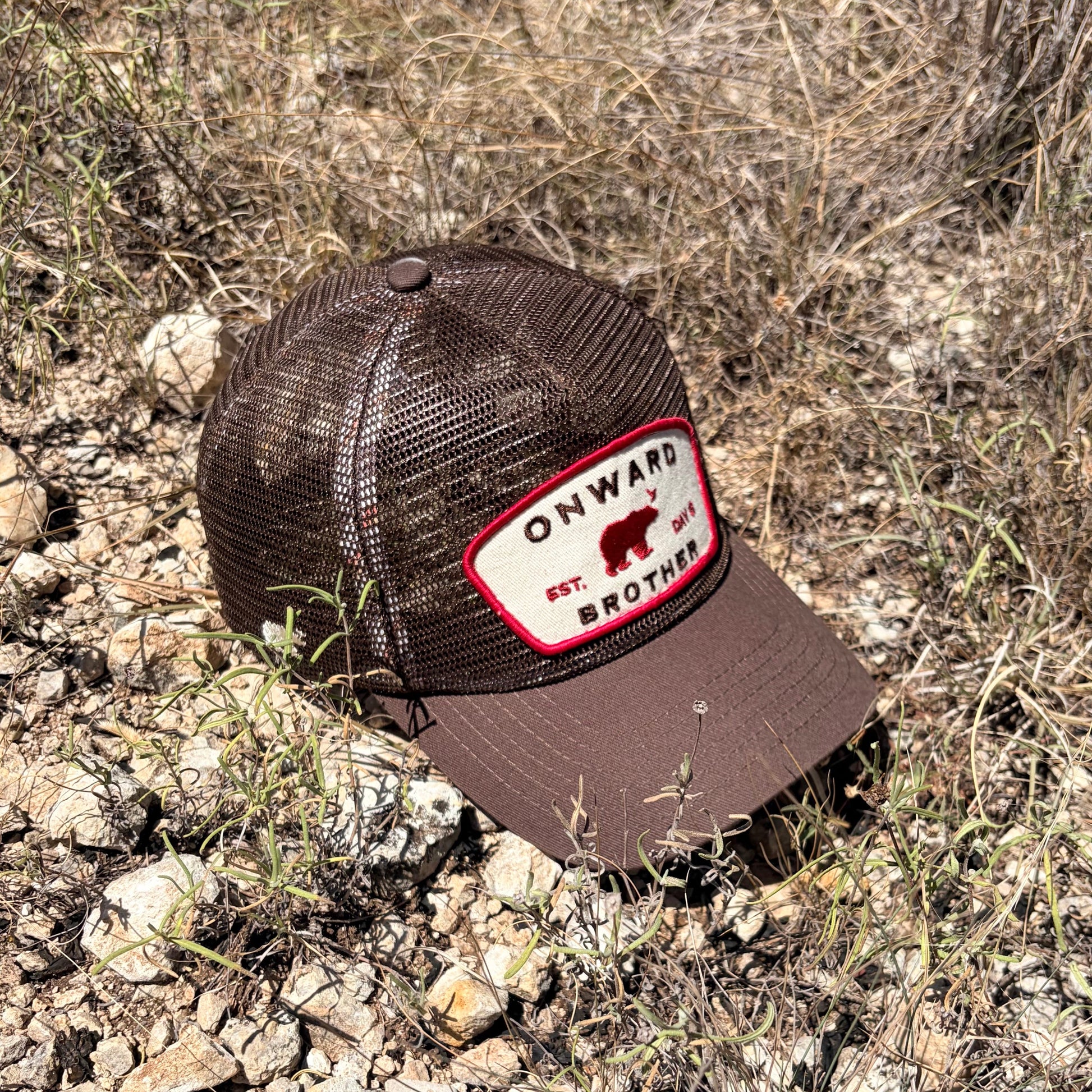 A brown trucker hat with a mesh back and a front patch reading 'ONWARD EST. DAY 6 BROTHER' lies on rocky, dry grass ground outdoors