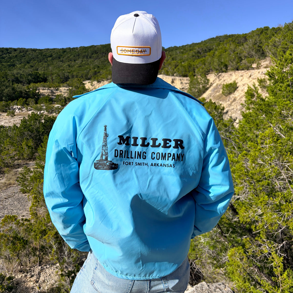 A man viewed from behind wearing a light blue windbreaker jacket with 'MILLER DRILLING COMPANY' printed on the back, standing outdoors in a rocky, green, and blue landscape. He's wearing the "someday" hat on backwards showing the word crossed out.