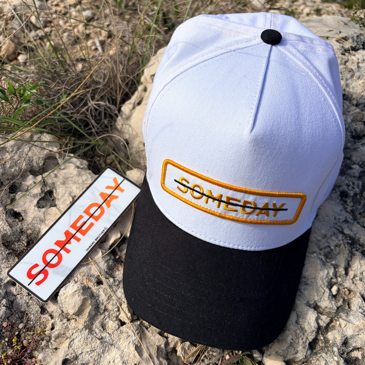 A white and black baseball cap with a yellow embroidered 'SOMEDAY' logo on the front, displayed on a rocky outdoor surface next to a matching hangtag.