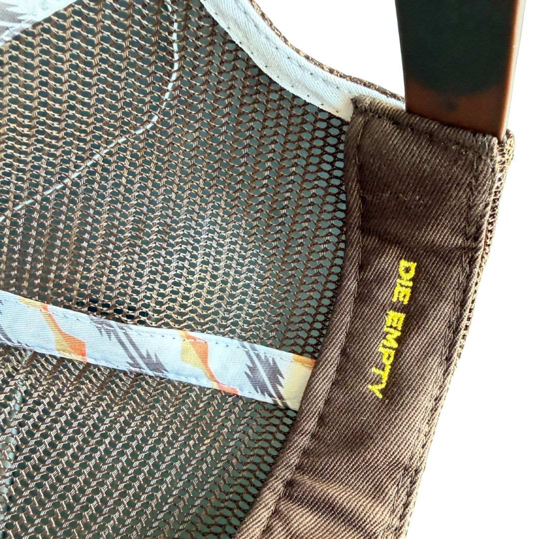 Close-up detail of a brown mesh hat showing the inner side, a patterned fabric band, and a brown fabric tab with yellow text reading 'DIE EMPTY'.