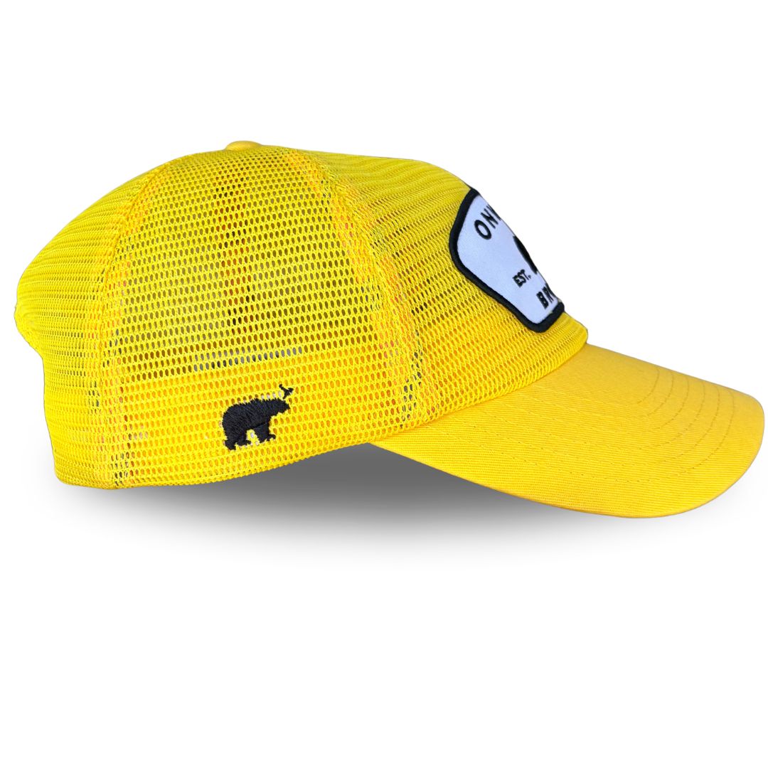 A bright yellow trucker hat with a white and black embroidered patch on the front that reads 'ONWARD BROTHER, est day 6' with a black bear silhouette. side view.