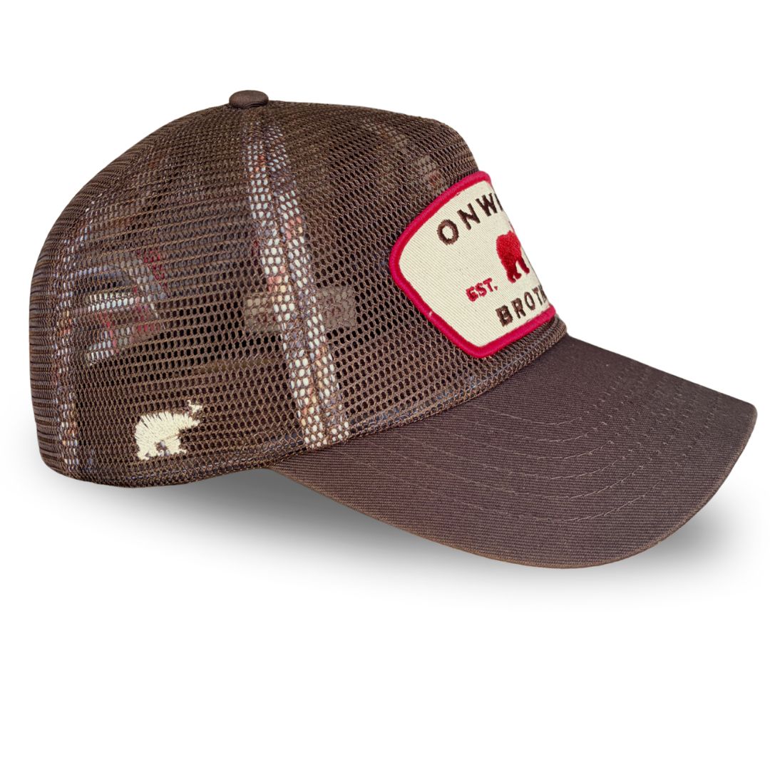 A brown trucker-style cap with a mesh back and a curved brim, featuring a cream-colored front patch with red trim and text that reads 'ONWILD EST. DAY 6 BROTHER' alongside a red bear logo.