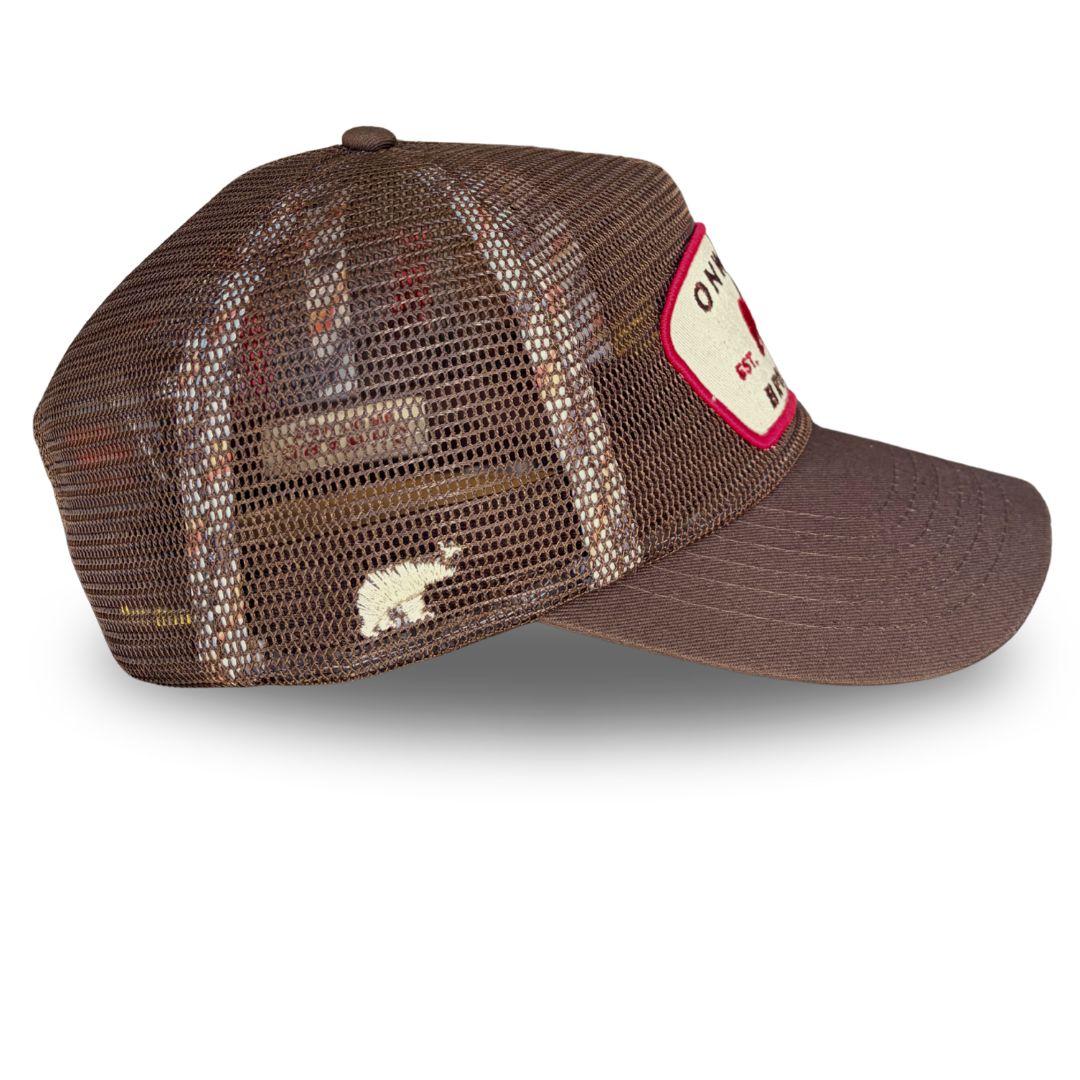 A brown trucker-style cap in all mesh mesh back, a brown visor, and a cream patch on the side with red trim.