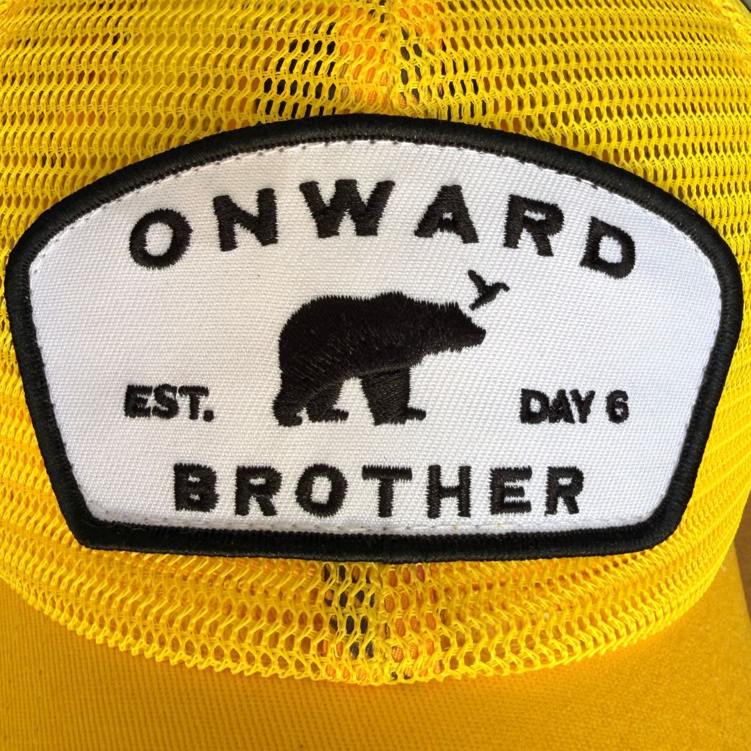 A close-up of a yellow trucker hat featuring a white embroidered patch with a black border. The patch displays the text 'ONWARD BROTHER', 'EST. day 6