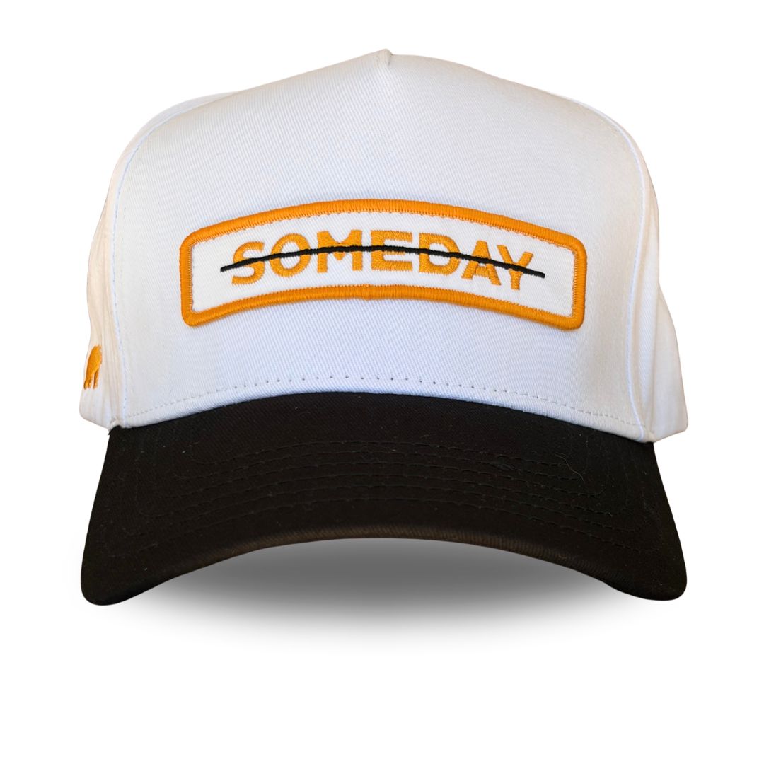 A white and black trucker-style cap with an orange and black embroidered patch on the front reading 'SOMEDAY' crossed out with a black line.