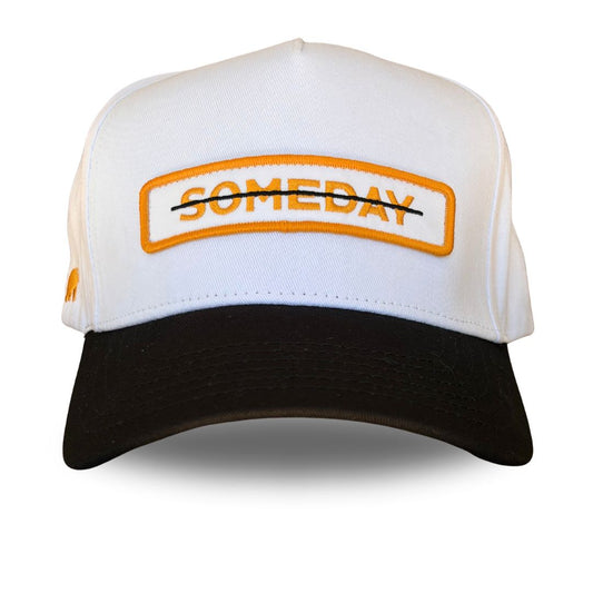 A white and black trucker-style cap with an orange and black embroidered patch on the front reading 'SOMEDAY' crossed out with a black line.