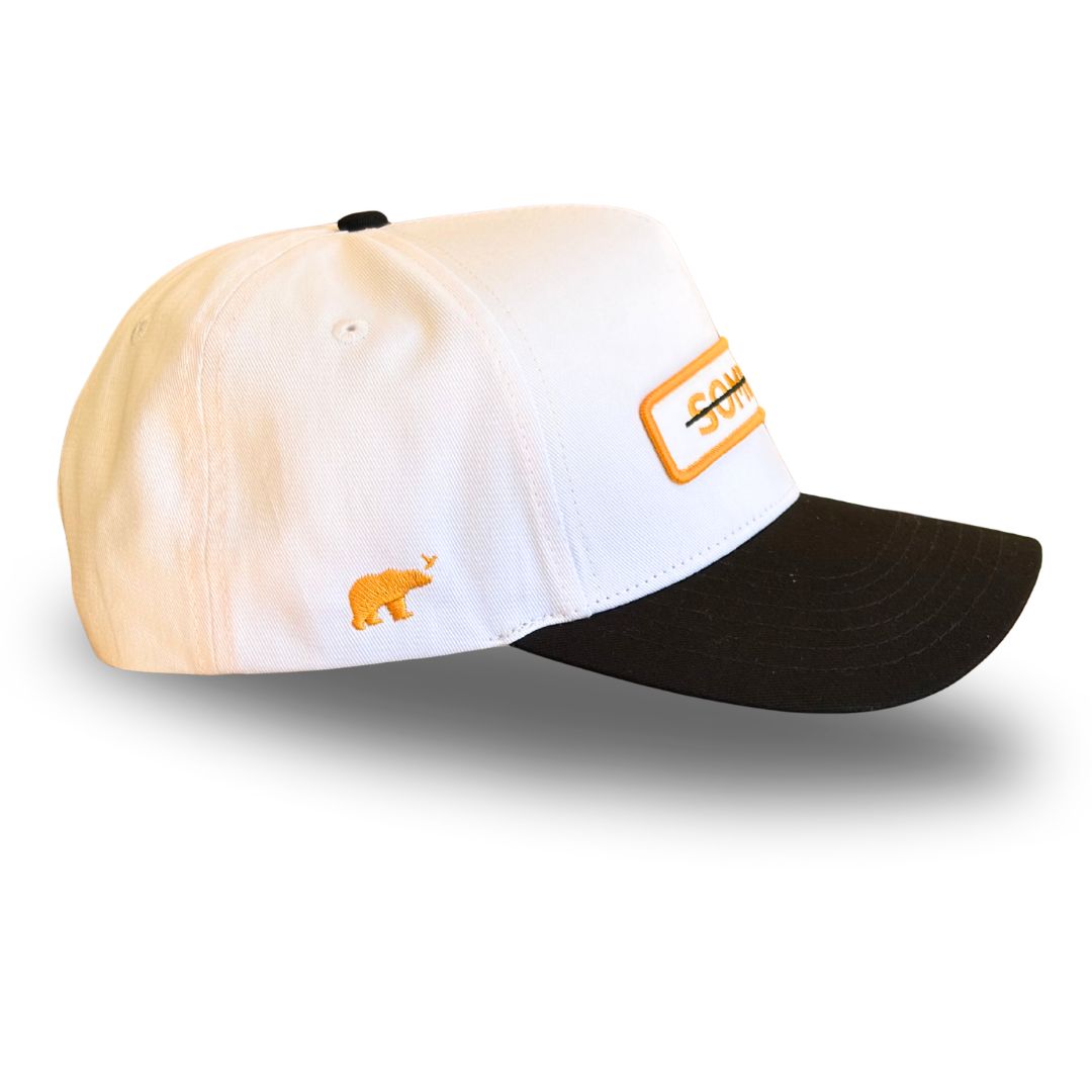 A baseball cap with a white crown, black brim, and orange embroidered details, including a small polar bear logo and a rectangular patch on the side.