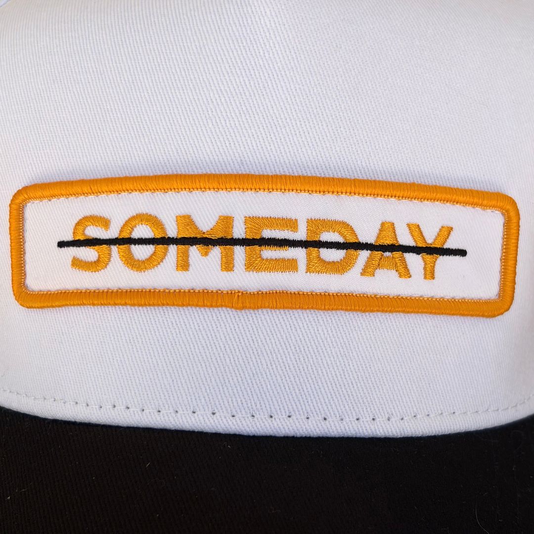 Close-up of a white baseball cap with a rectangular embroidered patch reading 'SOMEDAY' in orange, outlined in black, with the word crossed out with a black line.